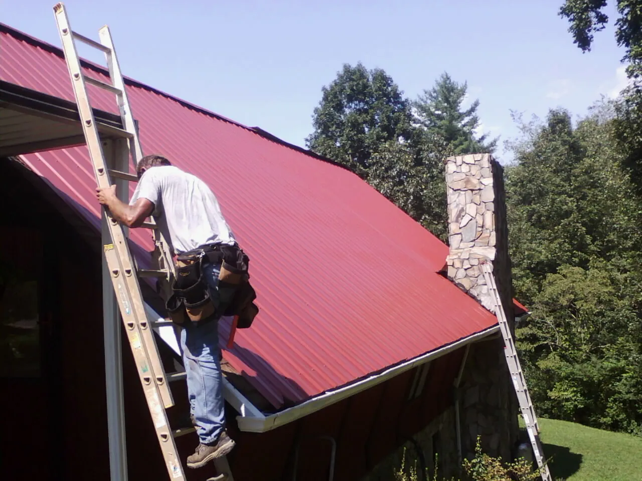 Roofer on a ladder beside a red metal roof and stone chimney — Soffit & Fascia Repair by Roofing Brothers in Williamson